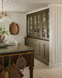 Beige glass-front kitchen cabinets with brass hardware beside a dark wood island; woven baskets hang on the wall above a light wood floor.
