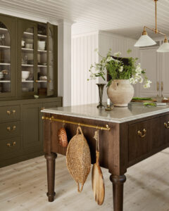 Dark wooden kitchen island with a marble countertop and brass hardware, topped by a large ceramic vase of white flowers; glass-front cabinets in the background.