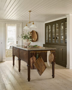 Bright kitchen with a dark wooden island, brass pendant light fixture, and glass-front cabinets against paneled walls with crown molding.