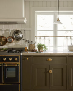 Dark kitchen with brass hardware, copper pots hanging on a rack above a gas range, white beadboard wall, and a bright window with a pendant light.