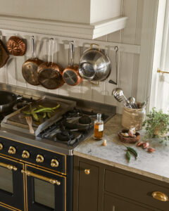 Copper and stainless steel pots hang on a rack above a gas stove in a modern kitchen; peppers, garlic, and a bottle of oil sit on the countertop.
