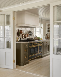 Professional kitchen featuring a black and brass double oven range, copper pots hanging on a rack, and a white range hood above the stove.
